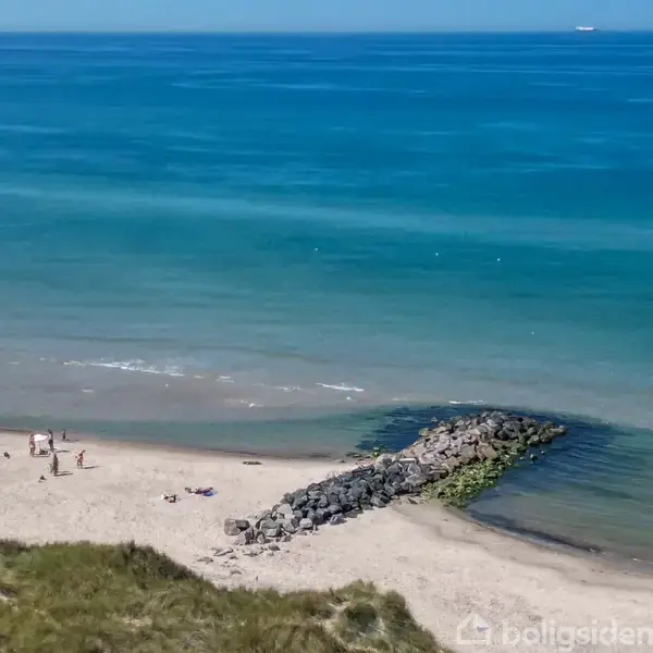 Mennesker solbader og går langs en sandstrand med klippefyldt mole, omgivet af roligt hav under en klar blå himmel.