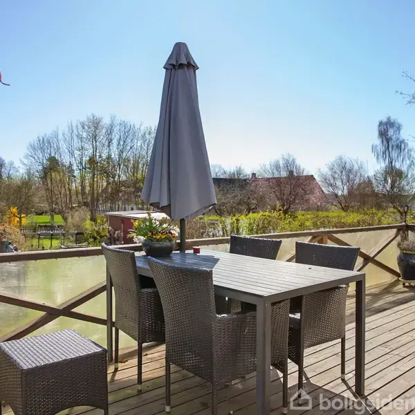 Terrassemøbler med parasol står på en hævet træterrasse i en have med udsigt til træer og en flagstang med Dannebrog. Solrigt vejr med blå himmel.