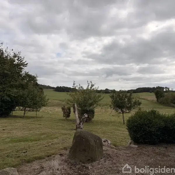 En stor sten står stille i en velholdt have med græsområder og spredte buske og træer, omgivet af en bakkefuld mark under en skyet himmel.