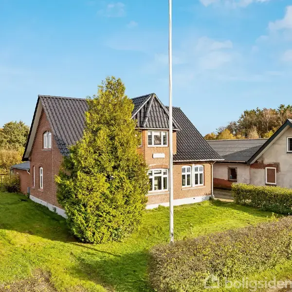 Et rødt murstenshus med sort tegltag står i en grøn have omgivet af træer og buske. Til højre ses en garage, og himlen er blå med enkelte skyer.