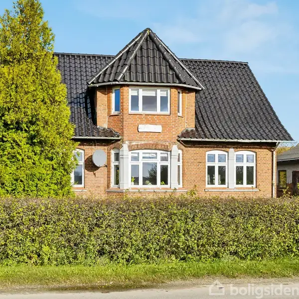 Rødt murstenshus med sorte tagplader, placeret bag en lav hæk. Foran huset ses en høj lygtepæl. Skyfri blå himmel bagved.