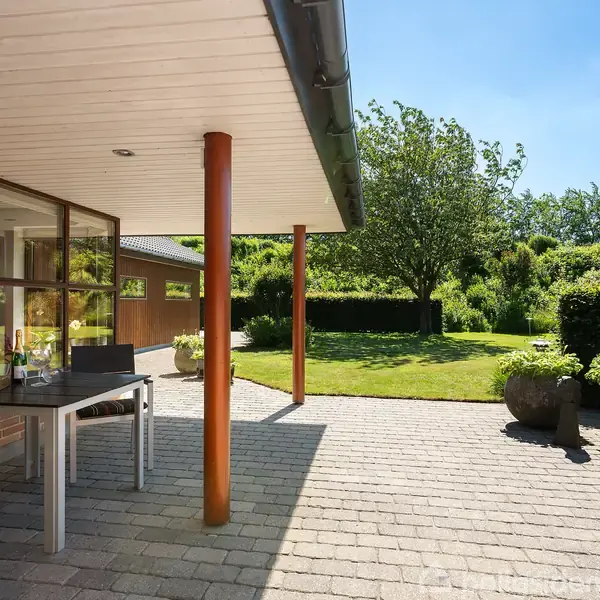 Terrasse med bord og stol står under husets tagudhæng. Græsplæne og træer i solrig have omringer terrassen. En flisebelagt belægning forbinder huset og haven.
