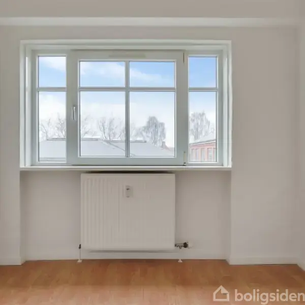 Et tomt rum med et stort vindue. Under vinduet er der en radiator. Udsigten viser snebeklædte træer og bygninger. Gulvet er af træ.