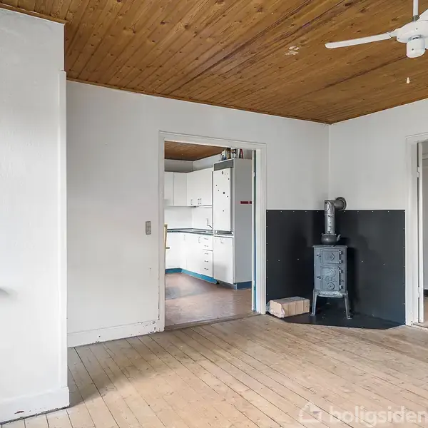 Et rum med trægulv og træloft, en brændeovn står i hjørnet. Der er en hvid loftsventilator. I baggrunden ses en lys køkkenindgang og en firkantet vinduesramme.