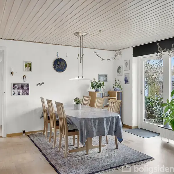Spisebord med seks stole står i et lyst rum med hvide vægge og trægulv. Der hænger billeder og en klokke på væggen. Store vinduer fører ud til en terrasse med planter.