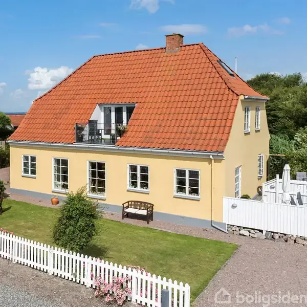 Gult hus med rødt tegltag står i en solrig have med hvidt stakit. Der er en terrasse med havemøbler, og omgivelserne inkluderer tætliggende hus og skovklædt baggrund.