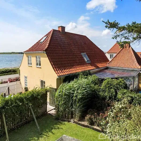 Et gult hus med rødt tegltag står ved vandet. Haven er frodig med buske og et lille hegn. En blå swimmingpool er synlig ved siden af huset.
