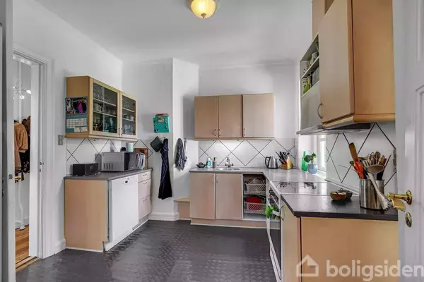 Køkken med træskabe og hvid flisevæg. Bordplader er dækket med køkkenredskaber og apparater. Mikrobølgeovn og opvaskemaskine til venstre. Hylder med glas og kopper ovenfor.