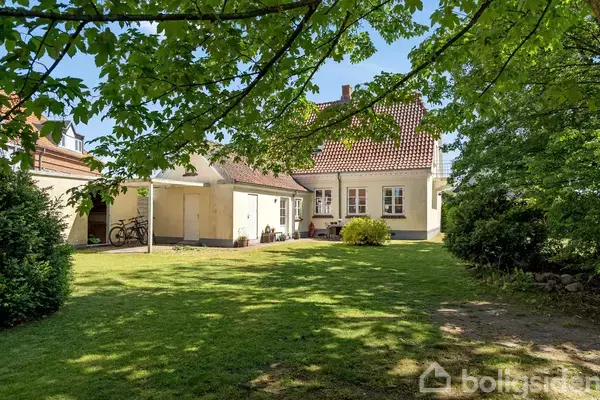 Hus med rødt tegltag står stille i solrigt haveområde, omgivet af græsplæne og træer. To cykler ses ved en tilbygning til venstre.