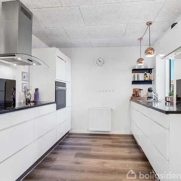 Et moderne køkken med hvide skabe og mørke bordplader. En frugtskål står på bordet. Loftet har hængende lamper, og der er en vægur på bagvæggen.