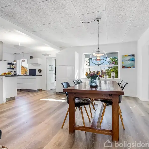 En stor spisebord med seks stole står i et lyst, åbent rum ved siden af et moderne køkken. Rummet har hvide vægge og billeder, med store vinduer mod haven.