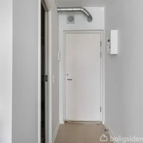 En hvid dør står for enden af en smal gang. Til venstre ses en del af et køkken med flaske på en bordplade. En ventilationsrør løber langs loftet.