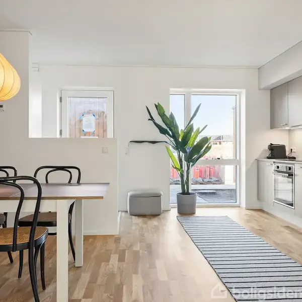 Spisebord med sorte stole står i en lyst køkken-alrum. En stor plante ved en altandør tilføjer grønne detaljer. Stribet tæppe løber langs køkkenet med grå skabe.
