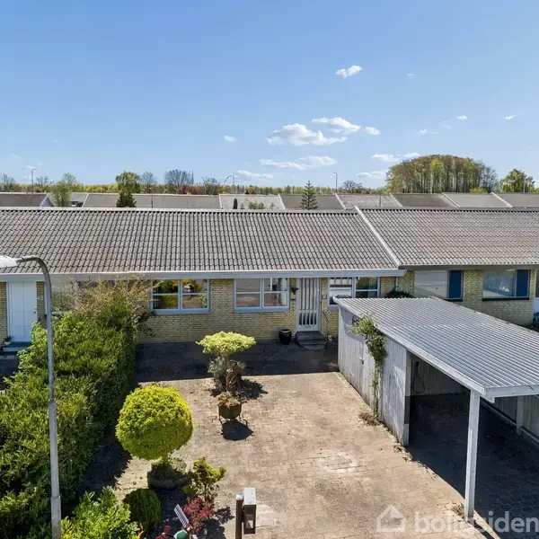 En rækkehus med carport står stille i et forstadsområde, omgivet af en velplejet have og en asfalteret indkørsel. Himlen er blå med få skyer i baggrunden.
