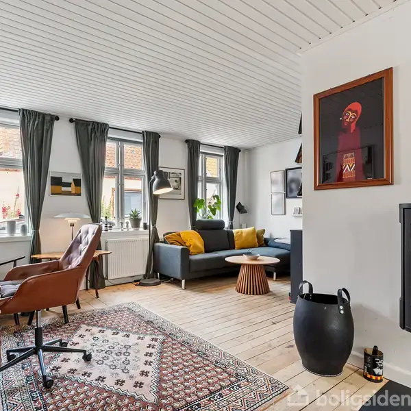 En stue med sofa og sofabord, indrettet med tæppe, lænestol ved skrivebord og pejs ved væggen. Vinduer med gardiner slipper lys ind; kunst hænger på væggen.