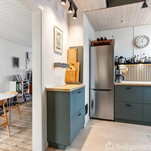 Køkken med grå skabe og køleskab til venstre, ur på væggen over kaffeudstyr. Bord med stole i tilstødende spisestue. Træloft og trægulv giver en hyggelig stemning.