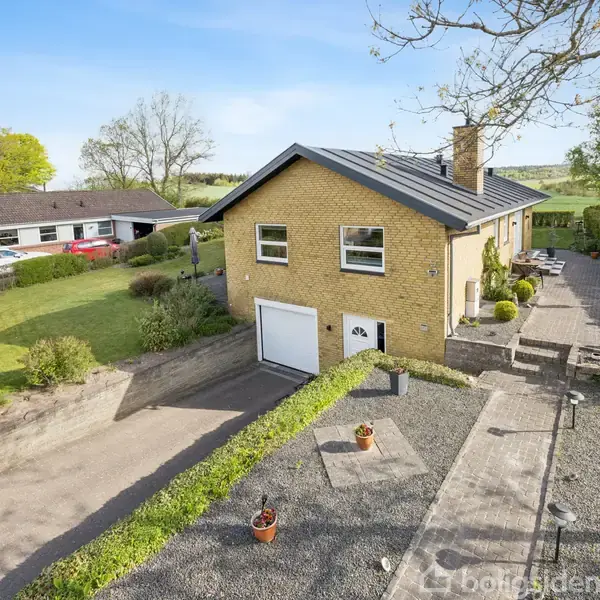 Et gult murstenshus med garage indbygget i kælderen ligger i en have med græsplæne og buske. Et flisebelagt terrasseområde løber langs siden af huset.