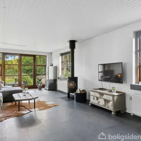 En stue med en grå sofa og sofabord står ved et vindue, der giver udsigt til en have. En brændeovn er placeret ved siden af et TV på en lav skænk.