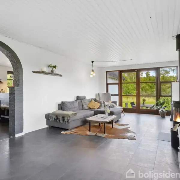 Stue med grå sofa og sofabord, ved siden af brændende brændeovn, i lyse omgivelser med store vinduer, der viser en grøn have udenfor. Åben bue fører til spisestue.