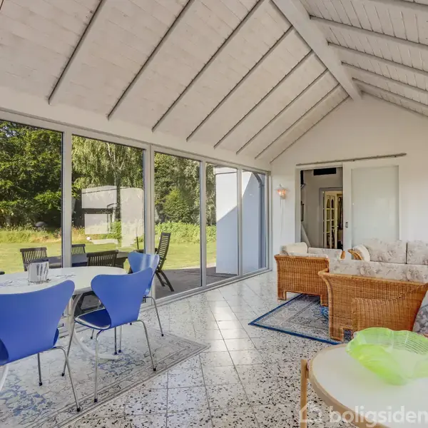 Spisestuemøbel med blå stole står i solskinsfyldt vinterhave med glasvægge. Sofaarrangement i rattan til venstre. Udsigt til terrasse og frodig have. Hvidt træloft og mønstret gulv.