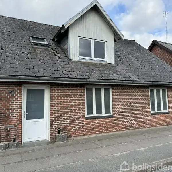 Rødt murstenshus med sort skifertag og frontkvist. Huset har en hvid hoveddør og flere vinduer. Det ligger ved en asfaltvej, der antyder et byområde.
