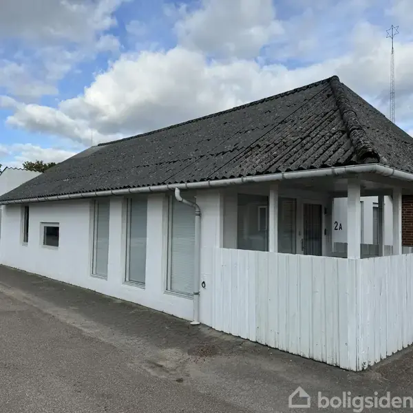 Hvidt hus med sort tag, placeret langs en vej. Nummer 2A er synligt på væggen. Bygningen har flere vinduer og en hvid træveranda under en delvist overskyet himmel.