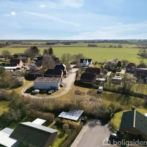 Boliger spredt i et landligt område med marker og træer i baggrunden. Et hvidt hus dominerer midten, omgivet af andre bygninger, mens veje forbinder strukturerne.