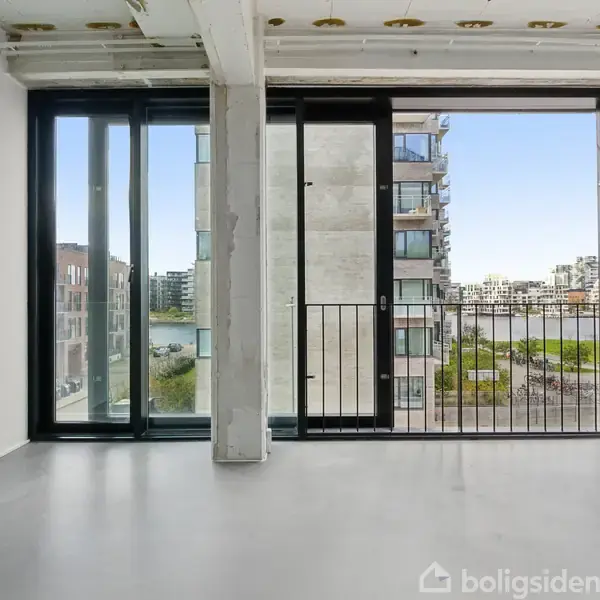 Stor glasdør åbner mod en balkon med udsigt over moderne bygninger og en vandkanal udenfor. Interiøret er lyst med hvide vægge og et gråt gulv.