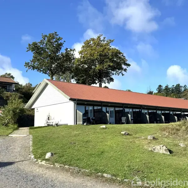 Lang bygning med rødt tag står stille i et grønt, åbent område med en grusvej og nogle træer i baggrunden under en blå himmel med enkelte skyer.