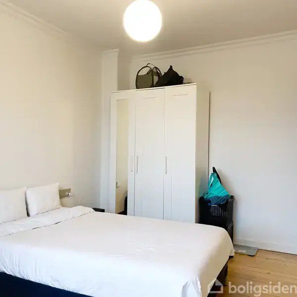 En seng med hvidt sengetøj står i et lyst værelse. En hvid garderobe ved siden af har tasker ovenpå. En dør er på højre side.