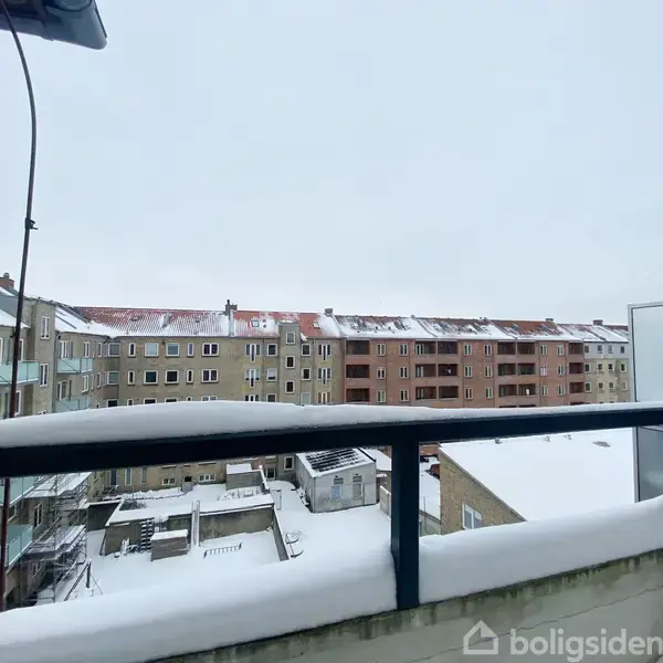 Balkonen er dækket af sne, mens den vender ud mod en række lejlighedsbygninger med snedækkede tage under en overskyet himmel.