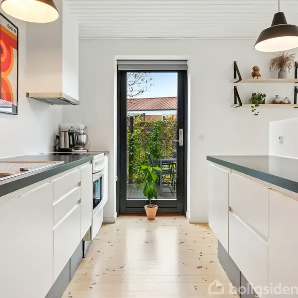 Et moderne køkken med hvide skabe og sort bordplade. En dør fører ud til en terrasse med planter. Hylder og lamper hænger på væggen. Malerier pryder rummets højre side.