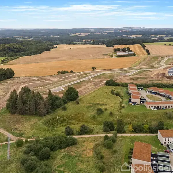 Landskab med marker og skovområder grænser op til en bebyggelse med røde taghuse. I baggrunden ses et landligt område med flerfarvede marker og byggeaktivitet.