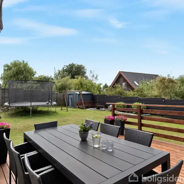 Et havebord med stole står på en terrasse. Bordet har planter som dekorationer. I baggrunden ses en have med trampolin, skur og hegn.