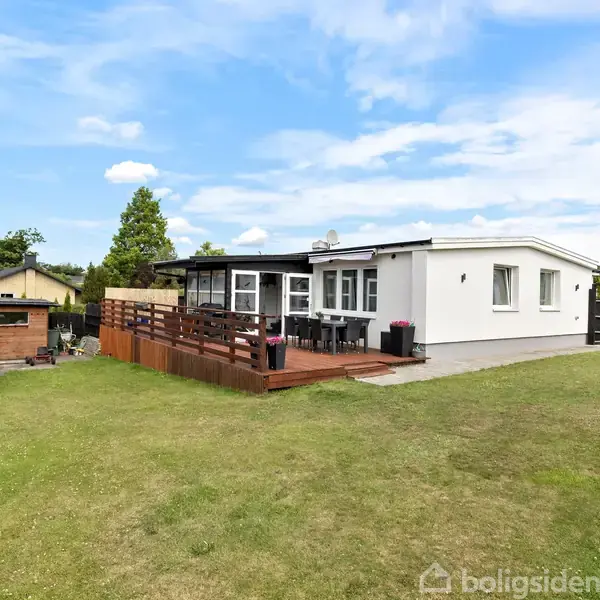 En hvid bungalow med store vinduer står stille. Den har en træterrasse med møbler i en velplejet have, omgivet af nogle små træhuse og grønne træer under en blå himmel.