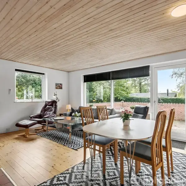 Stue med rundt spisebord, seks stole og lænestol ved glasdør til terrasse. Trægulv og lyse vægge. Vindue giver udsigt til have. Hylder med bøger og dekorationer langs væggen.