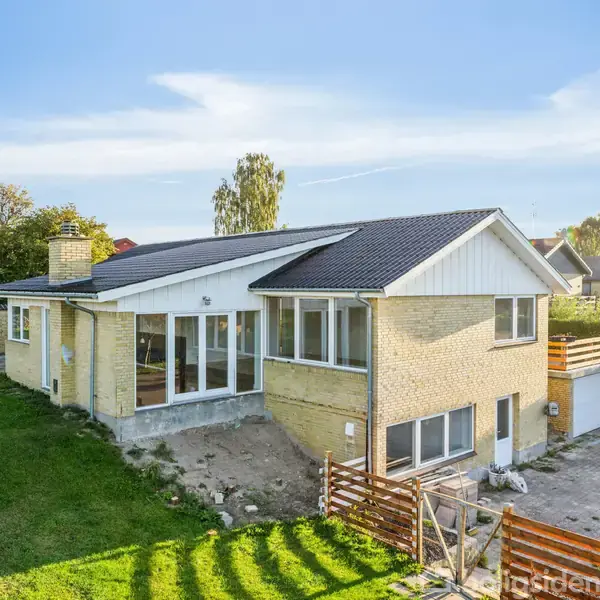 Huset har lyse mursten og et mørkt tag, ligger på en græsplæne med træomgivelser og har en terrasse med glaspartier. Der er en indkørsel på højre side.