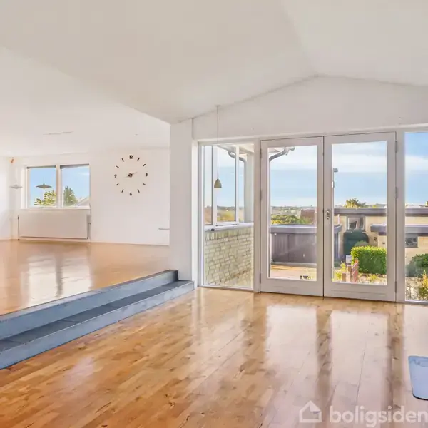 Stue med trægulv vender ud mod store vinduer, der giver udsigt til en have og havet. En ur hænger på væggen, og en brændeovn står i hjørnet.