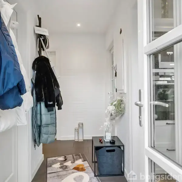 Gang med jakker hængende langs væggen, lys indretning. Til højre ses en dør med glasruder, og en kommode med dekorativ blomsteropsats. Tæppe på gulvet, småting arrangeret oppe.