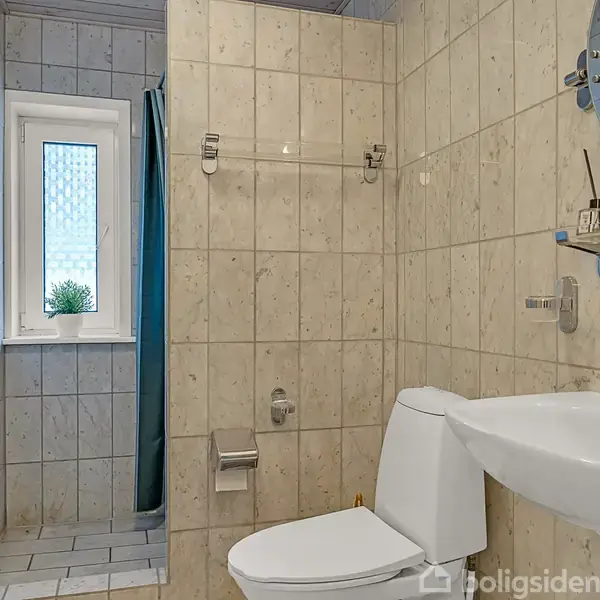 En ren, lys badeværelsesscene viser et toilet og en håndvask med spejl. Til venstre ses et vindue og en bruser med et blåt forhæng. Væggene er flisebelagte.