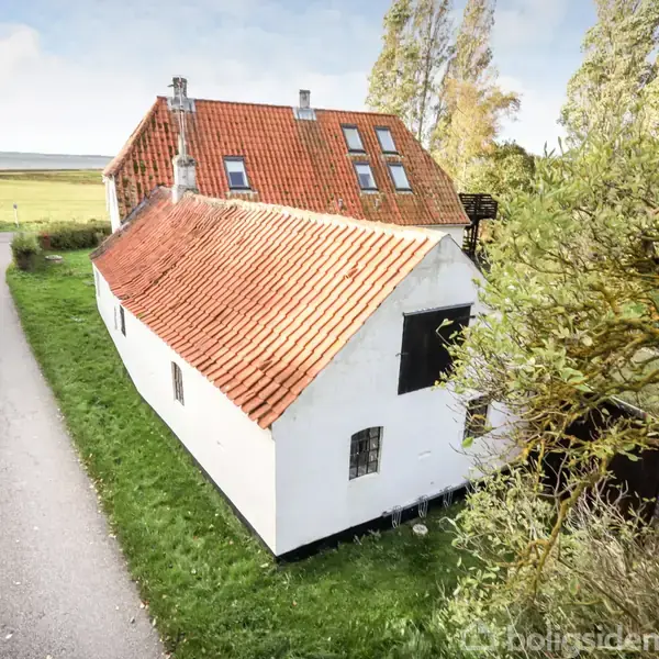 Hvidt hus med rødt tegltag står stille på en græsplæne ved en smal vej. Omkring huset er der frodig vegetation og åbne marker i baggrunden.