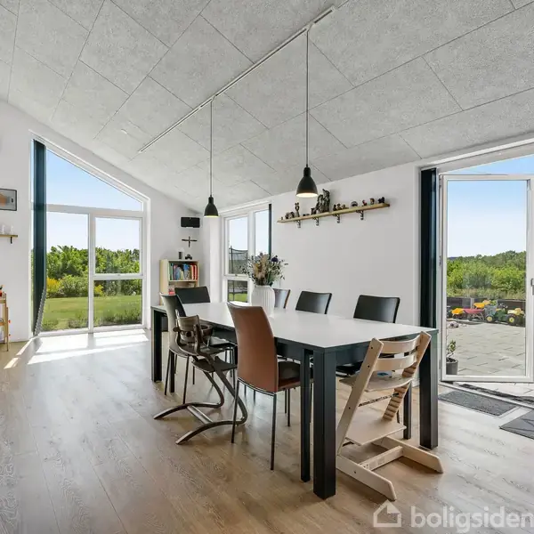 Et spisebord med stole står i centrum, oplyst af hængende lamper. Store vinduer og en dør åbner mod en terrasse og grøn udsigt, hvilket skaber en lys og åben atmosfære.