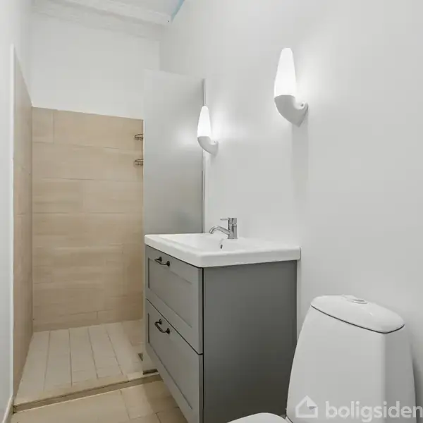 Badeværelse med et håndvaskskab og toilet i forgrunden, en bruseniche til venstre, og væglamper over håndvasken. Lyse vægge og enkel indretning præger rummet.