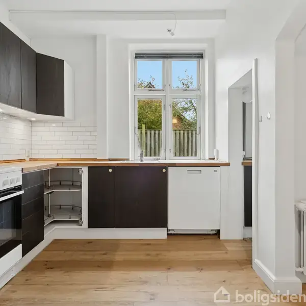 Et moderne køkken med sorte skabe og træbordplader, hvor der er en åben ovn og hvide fliser på væggen. Et vindue lader naturligt lys strømme ind.