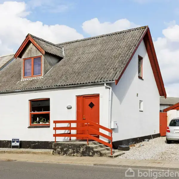 Et hvidt hus med røde døre og vinduesrammer står ved en vej. En hvid bil holder i gruset ved siden af huset. En skiltning ses foran.
