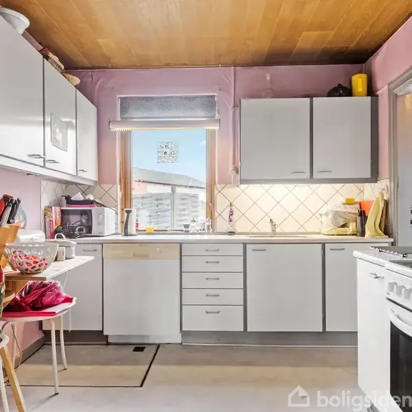 Køkken med hvide skabe og apparater står stille i et rum med lyserøde vægge og trægulv. Et vindue centralt giver naturligt lys. Bordpladerne er delvist optaget af køkkenudstyr.