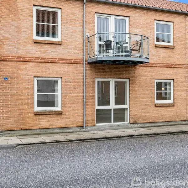 To-etagers murstensbygning med balkon. På balkonen står bord og stole. Bygningen har flere vinduer. Beliggende ved fortov og vej. Adressenummeret 82 er synligt ved døren.