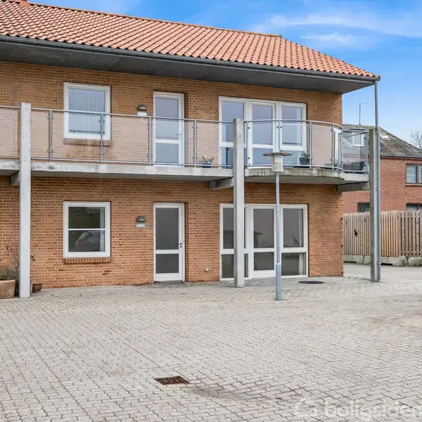 Rødstensbygning med to etager; øverst har balkon med glasrækværk. Foran bygningen ses en brolagt gårdsplads; til venstre står et lille bord med stole og planter.
