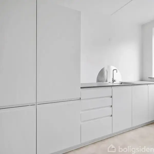 Et moderne køkken med hvide skabe står stille i et lyst rum. Der er en bordplade med en indbygget vask og vindue med udsigt til en by.