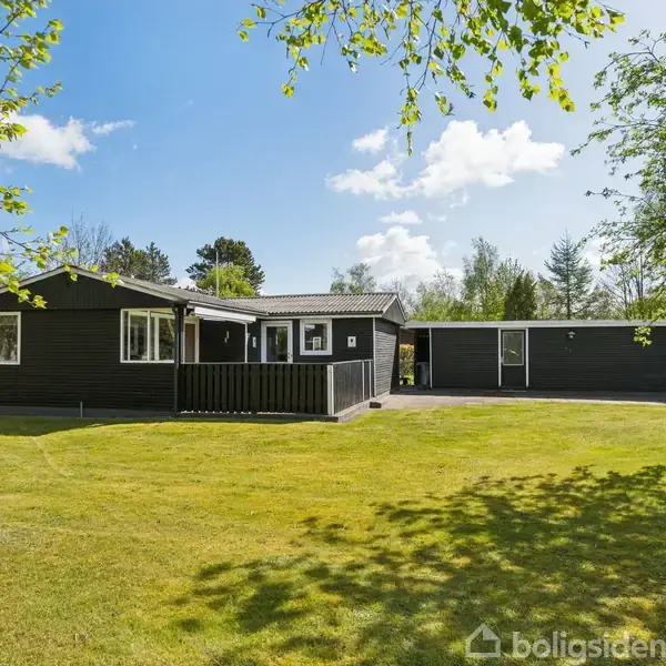 Sort træhus står på en stor græsplæne. Flere træer omgiver ejendommen, og himlen er klar med nogle få skyer.